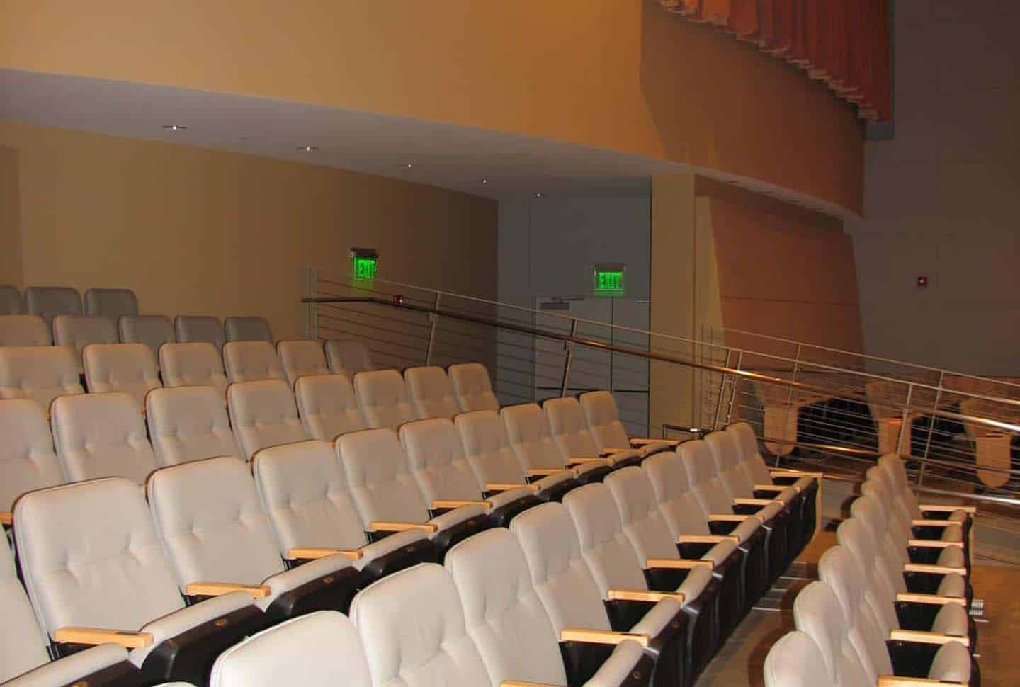 Rows of empty beige theater seats face a stage in a modern auditorium, with green exit signs and a metal railing visible in the background.
