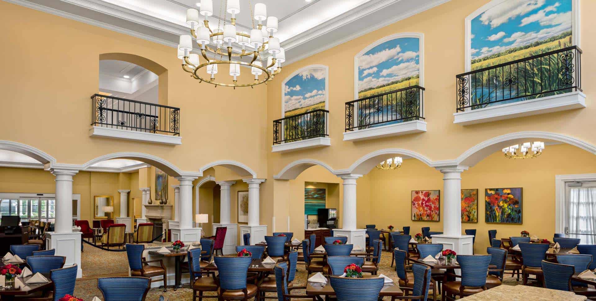 A spacious dining area with blue chairs, white columns, chandeliers, large landscape paintings, and tall windows under a high ceiling.