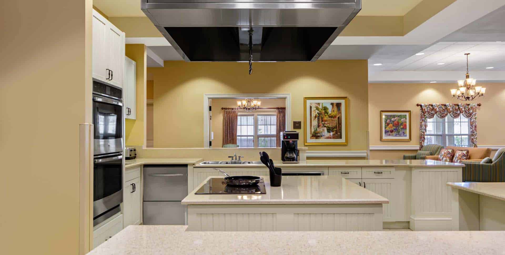 Modern kitchen with beige walls, white cabinets, stainless steel appliances, and an electric cooktop on a large island; paintings and light fixtures in the background.