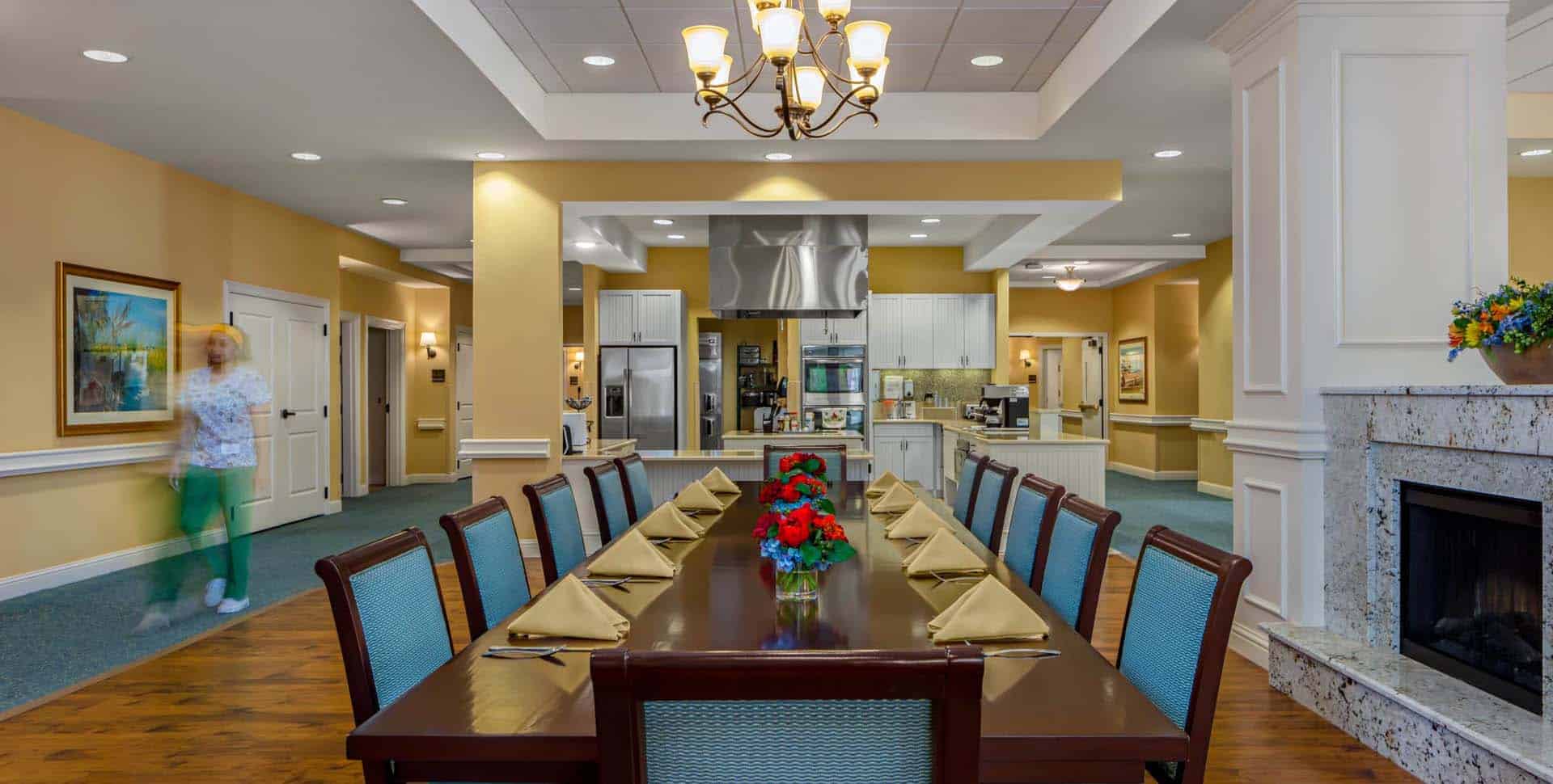A dining room with a long table set for ten, yellow napkins, red flower centerpieces, an open kitchen, and a blurred person walking in the background.