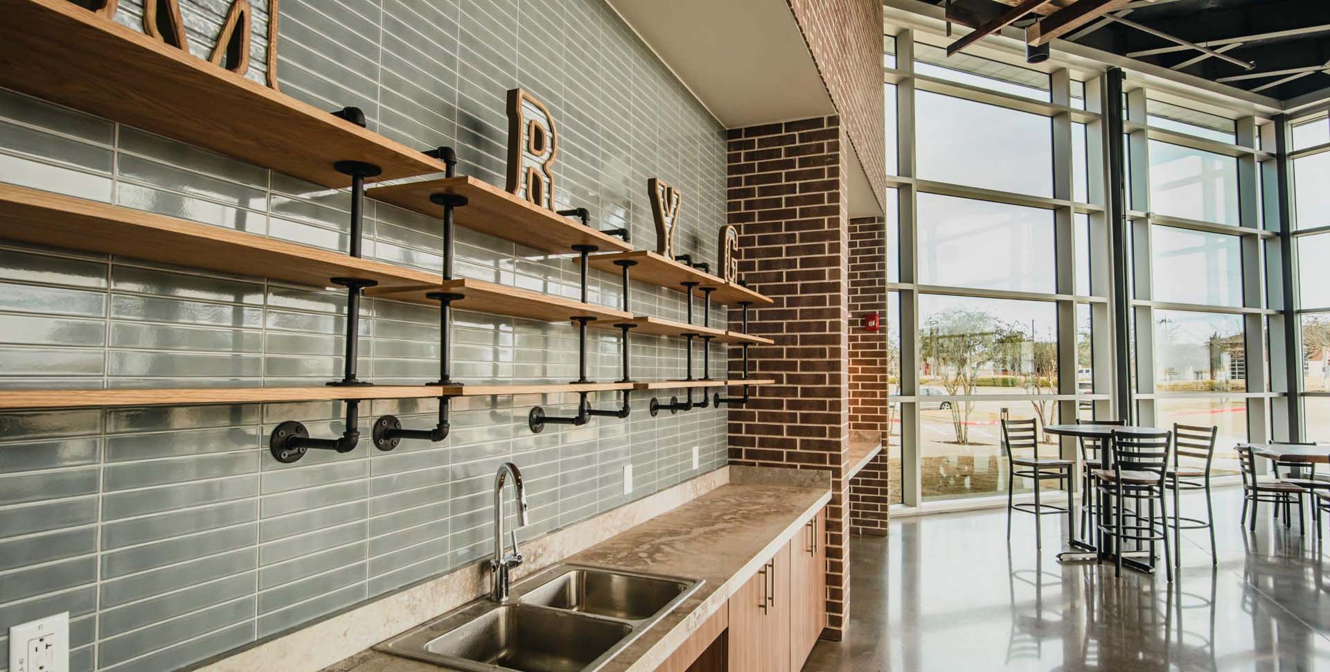 Modern kitchen area with a stainless steel sink, open wooden shelves supported by black metal pipes, brick and tile walls, and large windows overlooking an outdoor seating area.