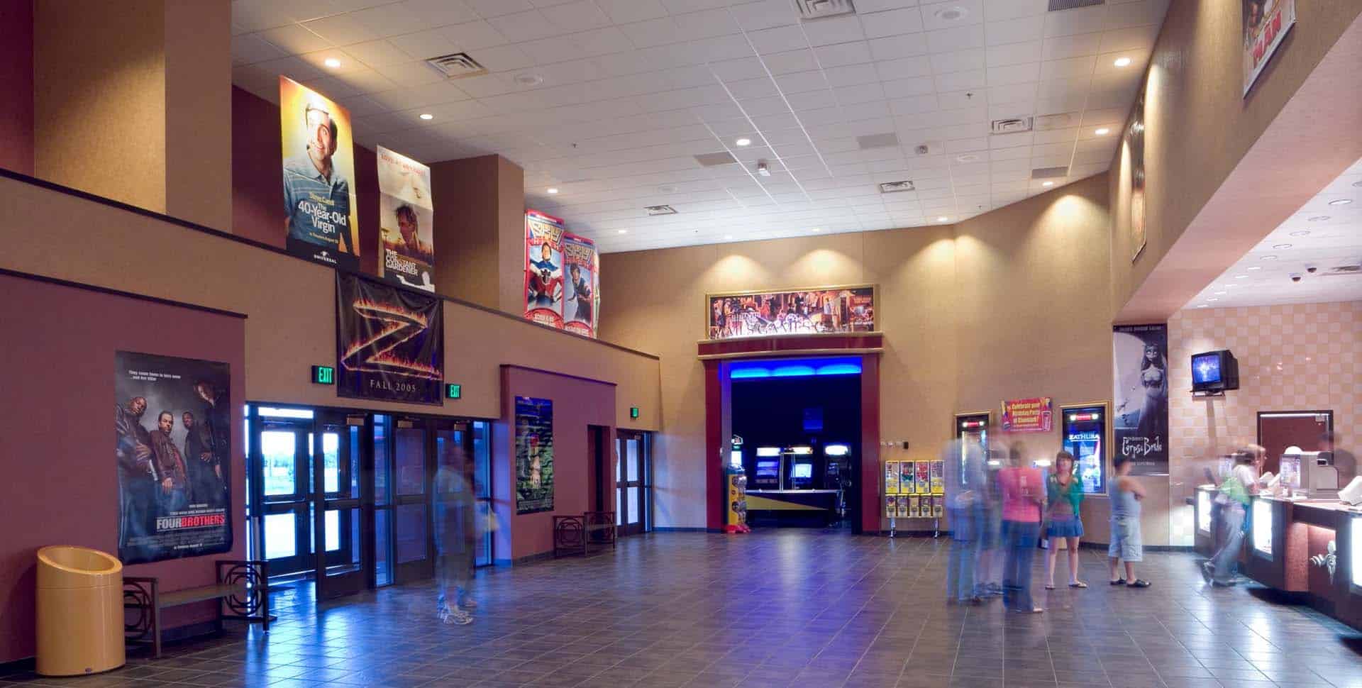 Spacious movie theater lobby with posters on the walls, people standing near ticket counters, vending machines, and a snack bar visible on the right.