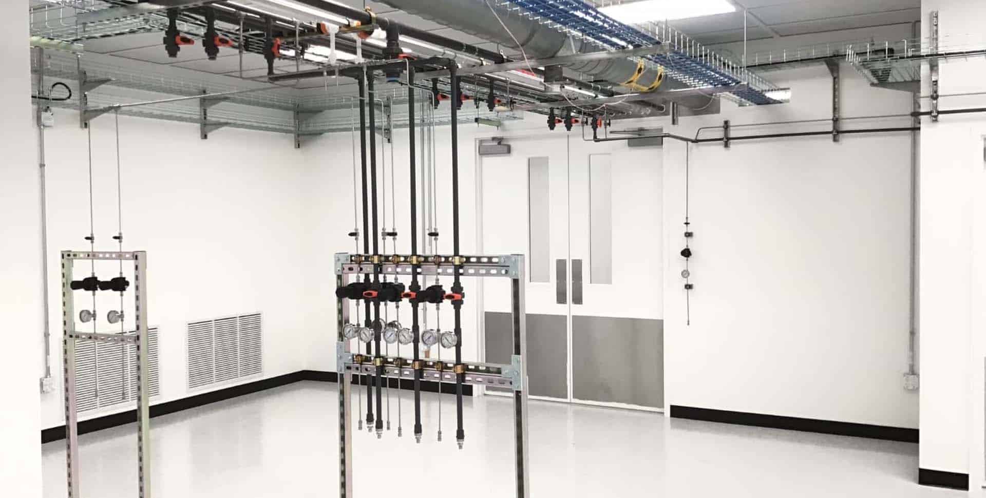 A clean, white industrial room with exposed pipes, valves, ceiling cables, ventilation panels, and glass double doors in the background.