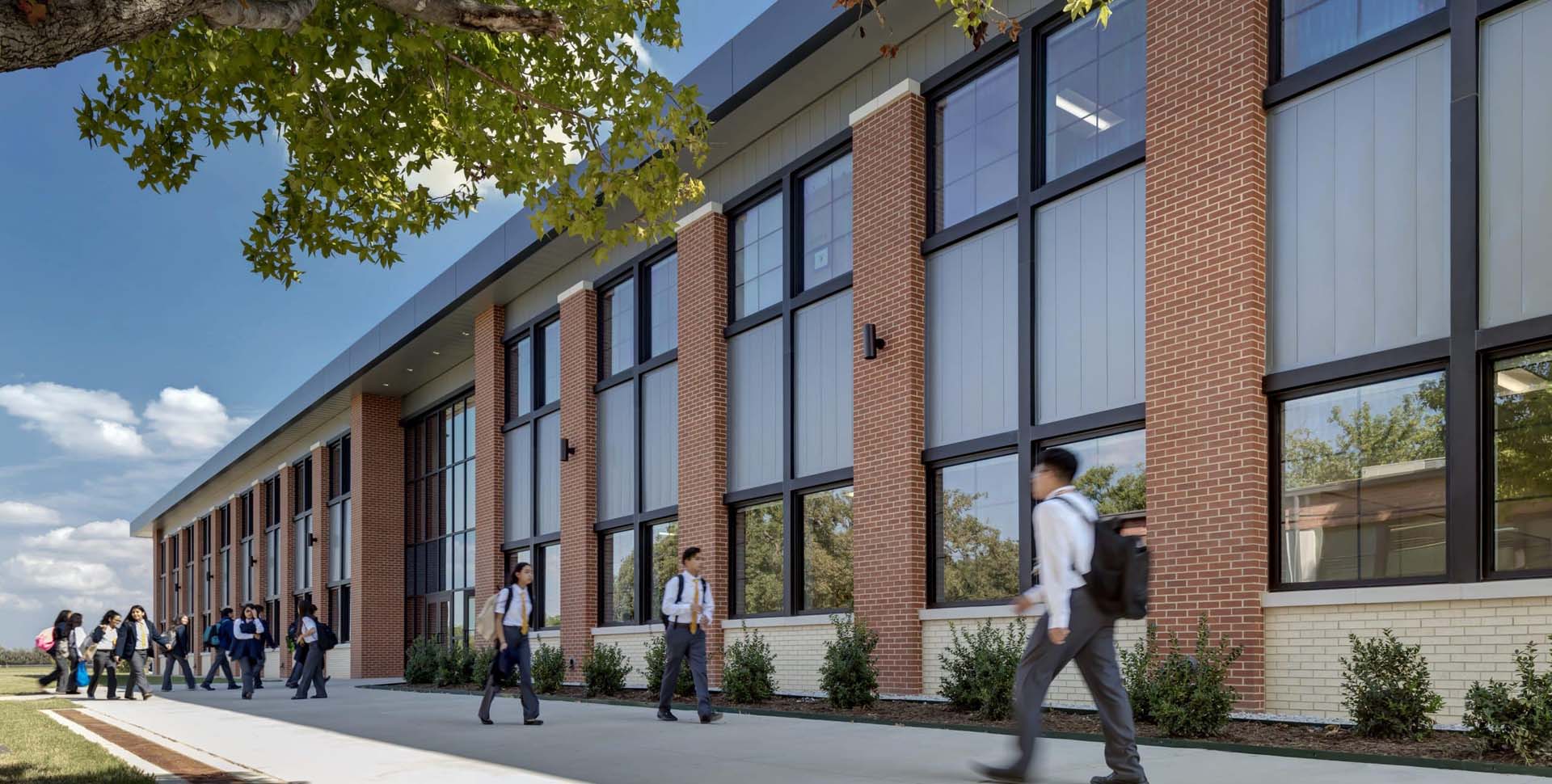 Students walk on a sidewalk outside a modern brick school building with large windows on a sunny day.