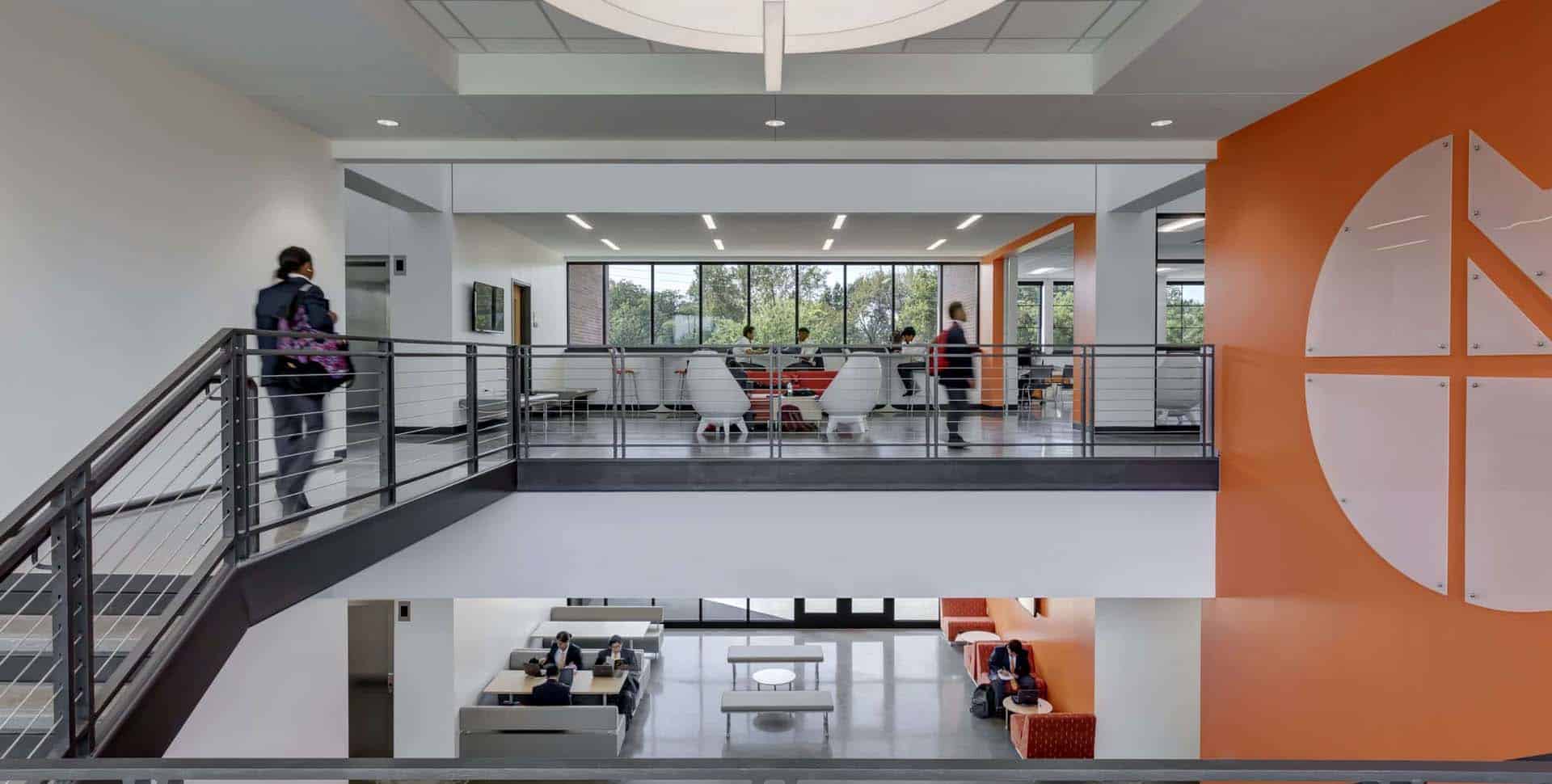 Modern office or school interior with people walking and sitting; open layout features glass railings, orange accent wall, and natural light from large windows.