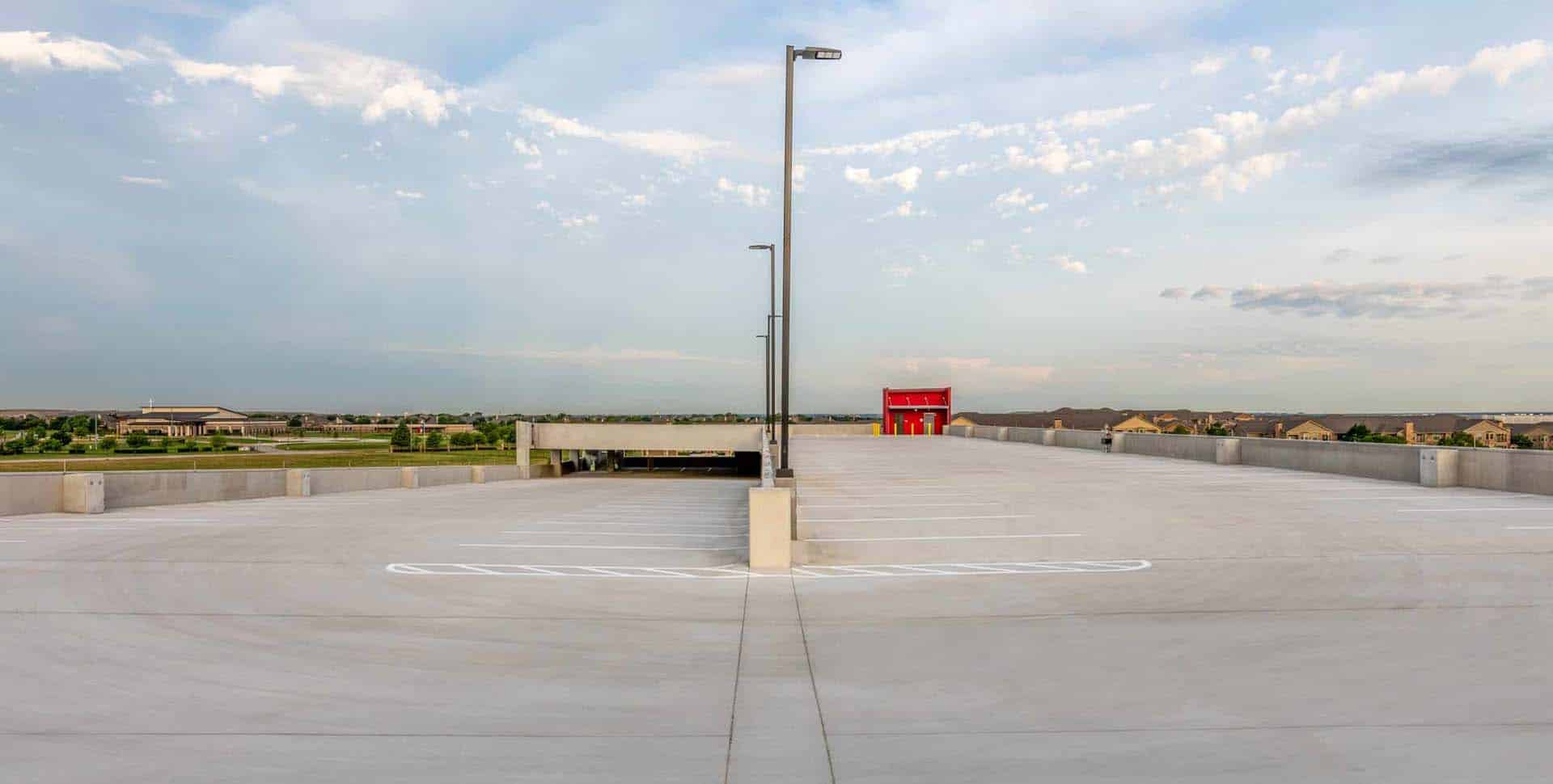 Empty rooftop parking lot with a central street lamp, white parking lines, and a red stairwell structure in the background under a partly cloudy sky.