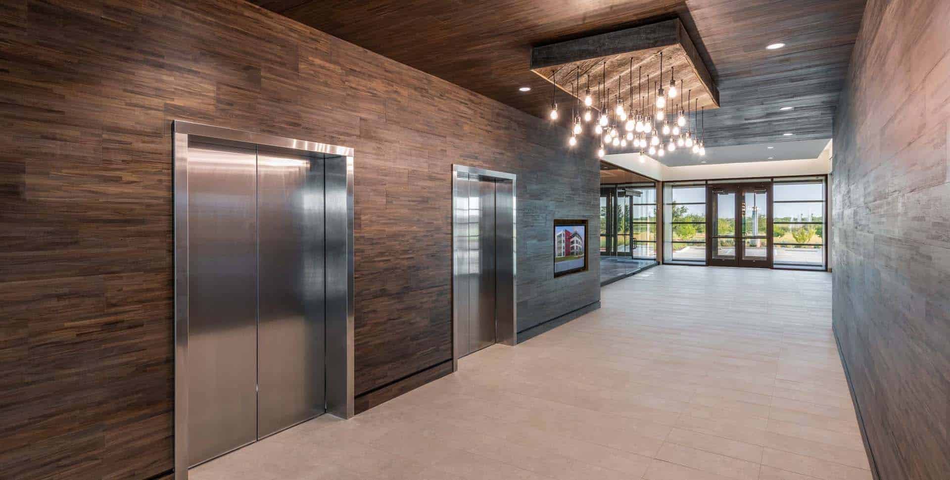 Modern building lobby with two stainless steel elevators, wood-paneled walls, tile flooring, and a chandelier, leading to glass entrance doors with an outside view.