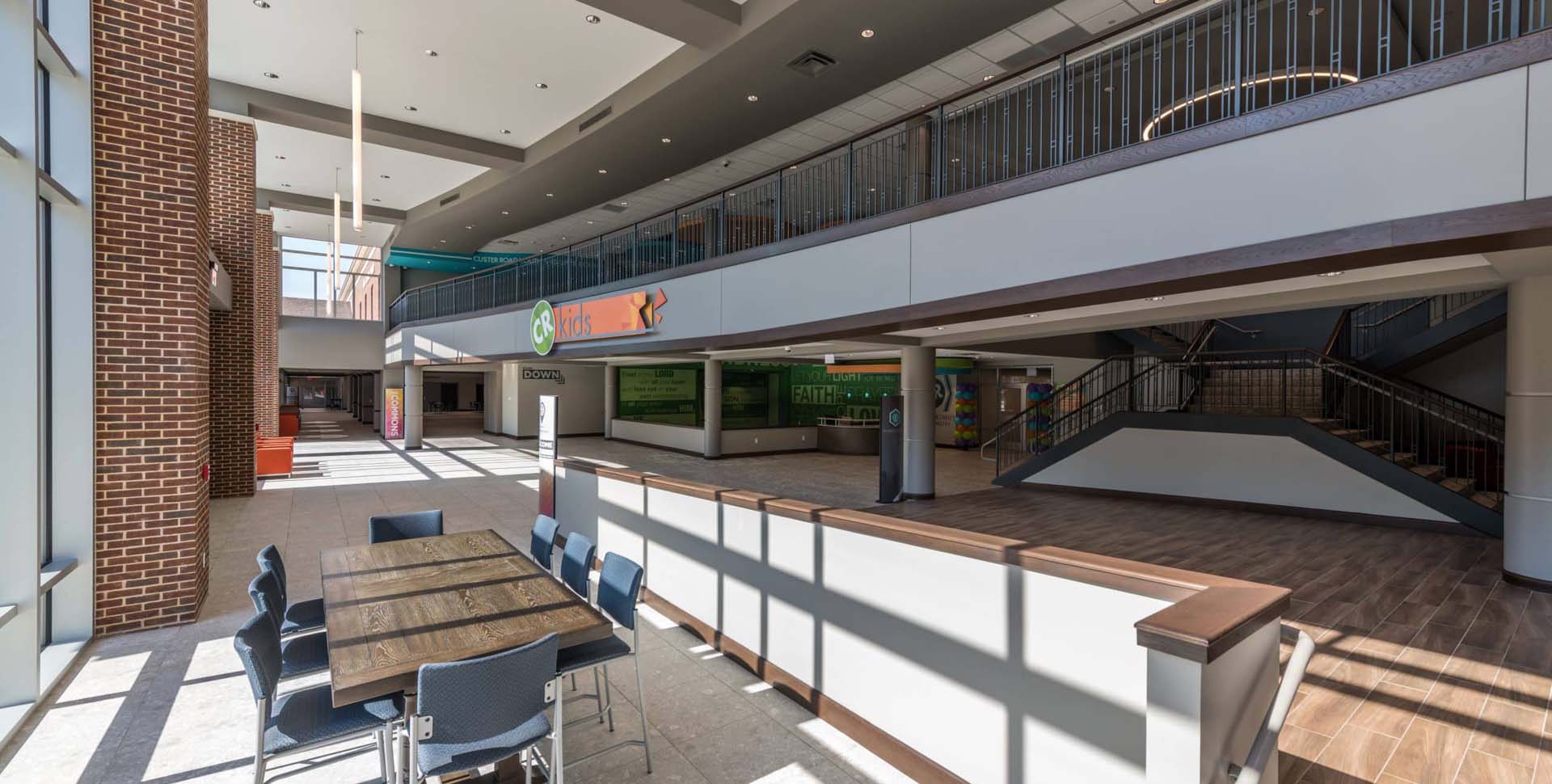 A spacious, modern lobby with tables and chairs, large windows, a staircase, and second-floor railing in a commercial or educational building.