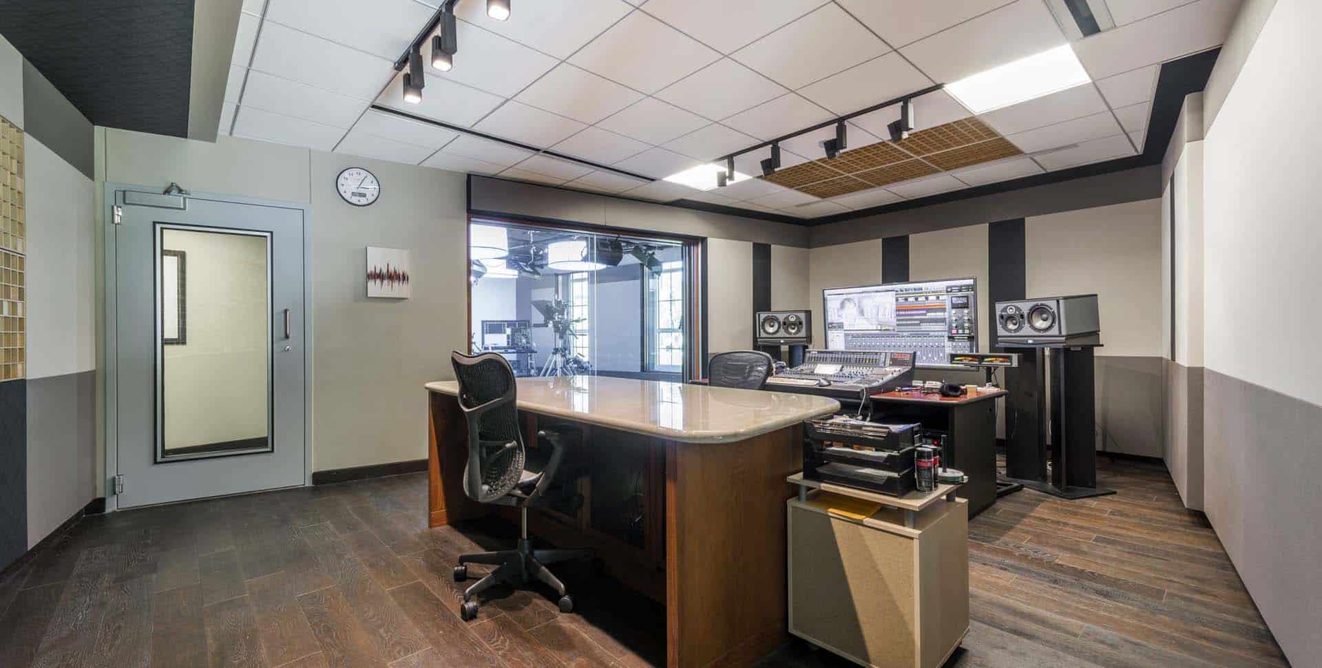 Modern recording studio with a mixing desk, two chairs, studio monitors, a computer, and a large window looking into an adjacent room.