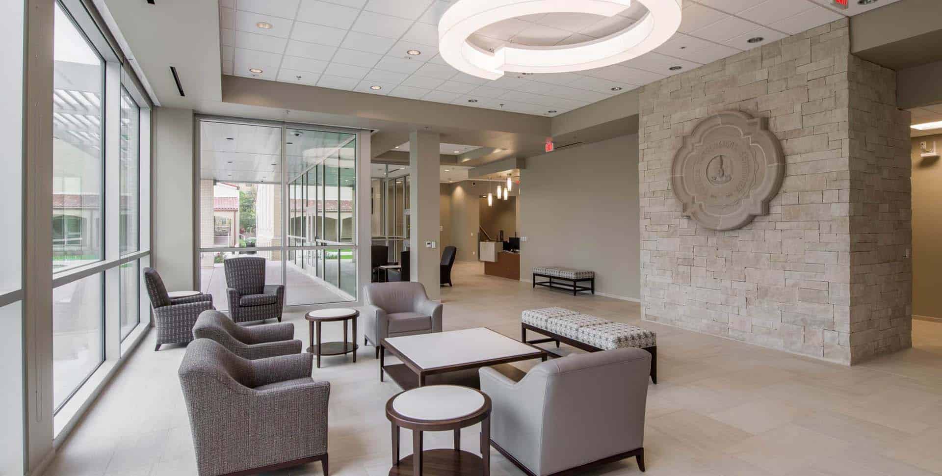 Modern lobby with gray armchairs, round tables, and a stone accent wall. Large windows and a circular ceiling light create an open, well-lit space. Reception desk visible in the background.