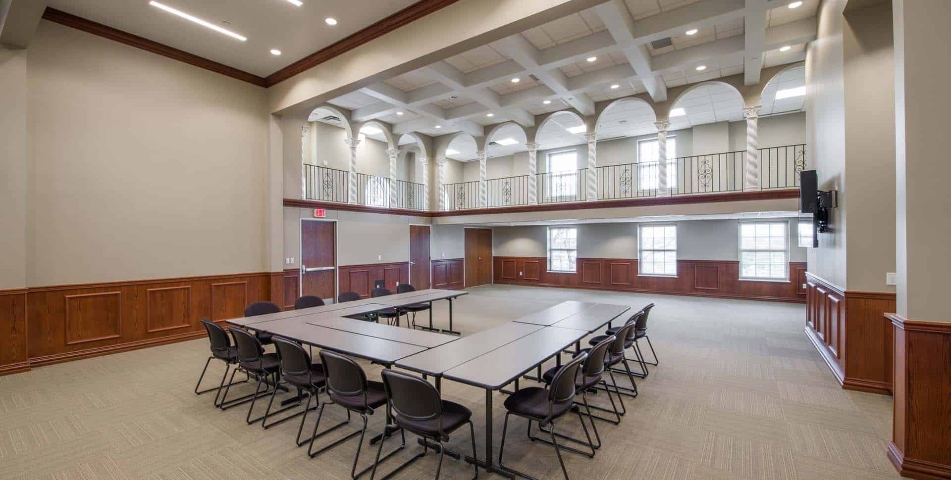 Spacious conference room with U-shaped table setup, black chairs, high ceiling, large windows, wooden paneling, and a balcony area.
