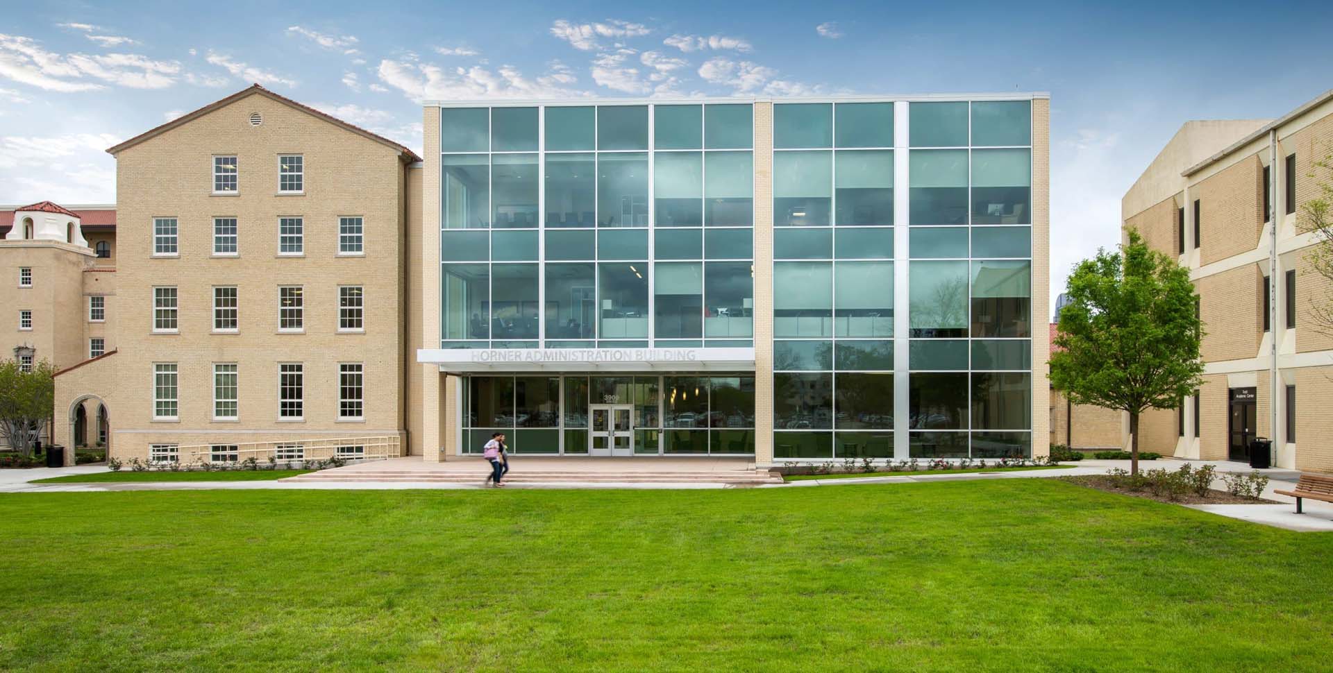 A modern glass-fronted building labeled "John & Anna Graves Hall" stands between two older brick buildings, with a person walking across the green lawn in front.