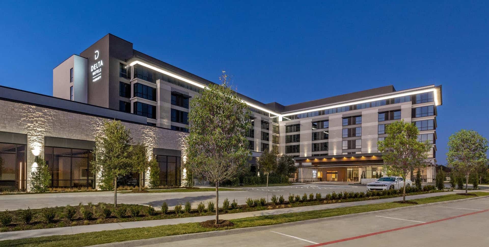 Delta Hotels by Marriott exterior at dusk with illuminated lights, landscaped entrance, and parked cars in front of a modern multi-story building.