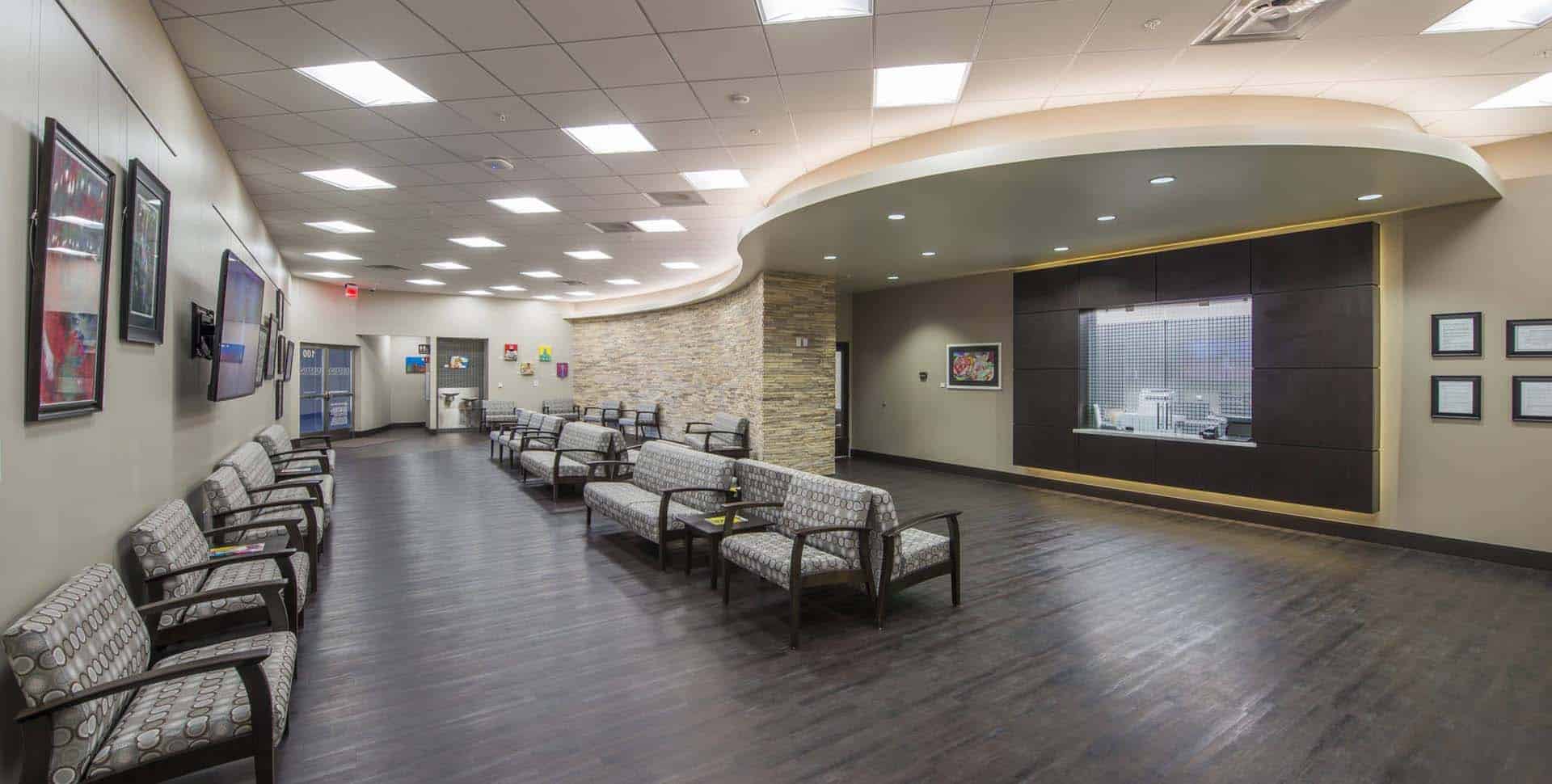 Modern medical waiting room with rows of patterned chairs, a reception window, framed art on the walls, and overhead fluorescent lighting.