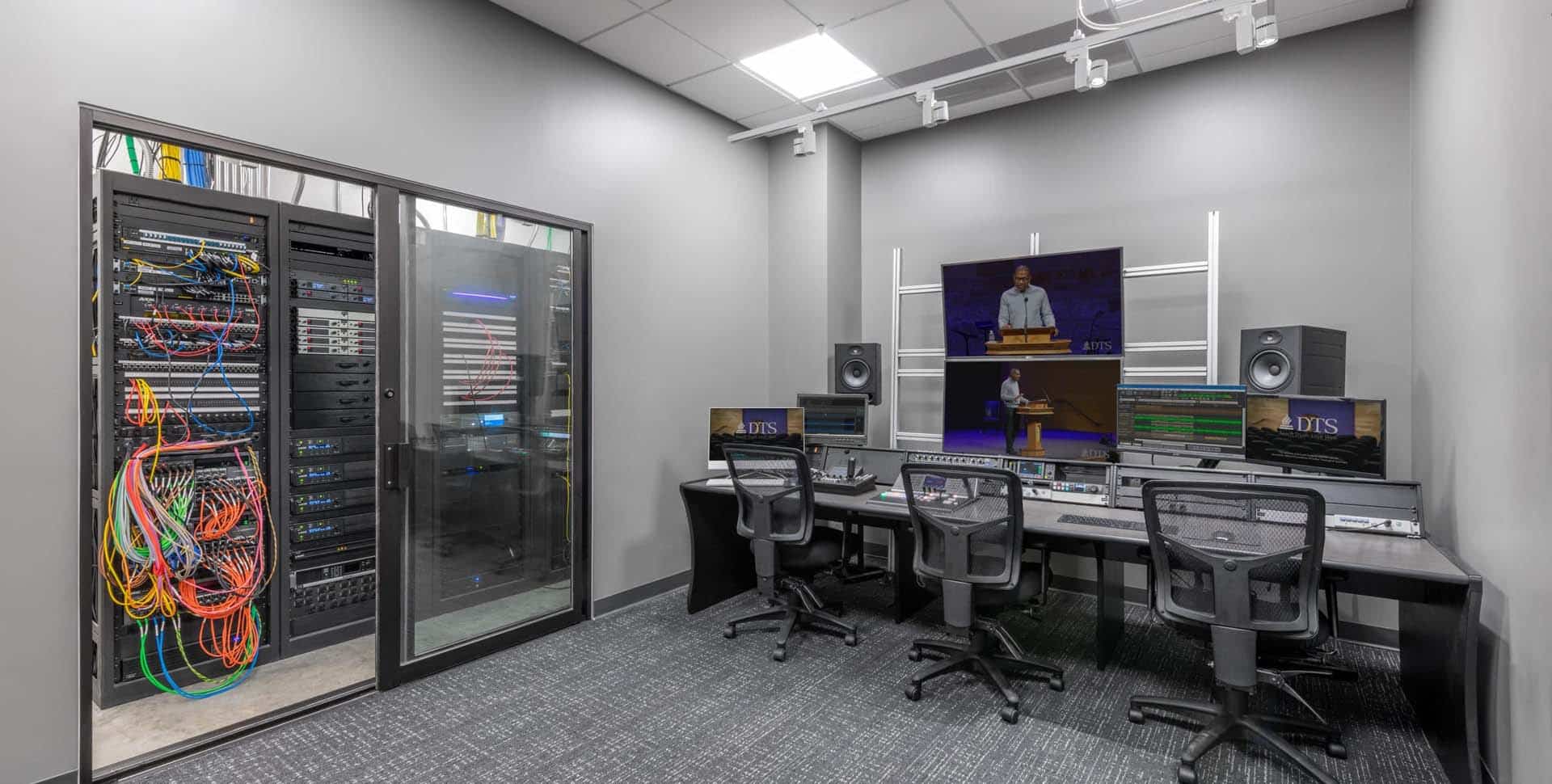 A modern control room with three workstations, computer monitors, large speakers, and a wall-mounted screen, next to a server room with network cables and equipment.