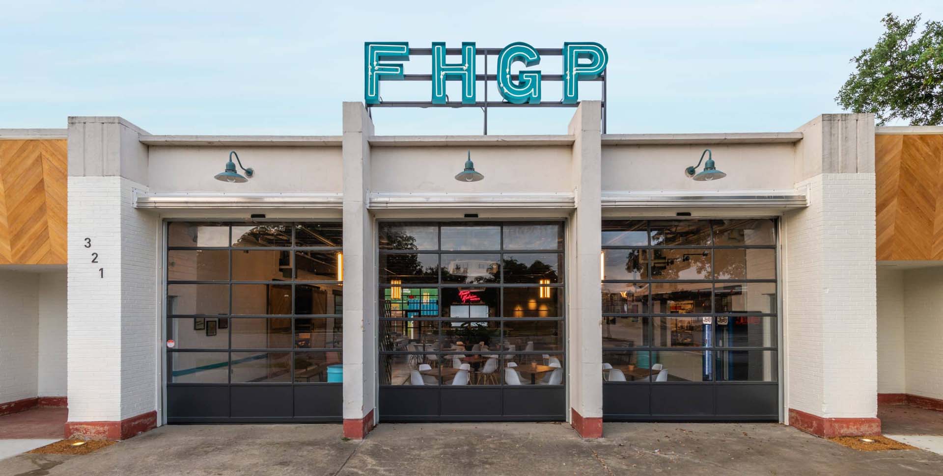 A building with large glass garage doors and a blue neon “FHGP” sign on the roof, with outdoor lights above each door and the street number 321 on the left side.