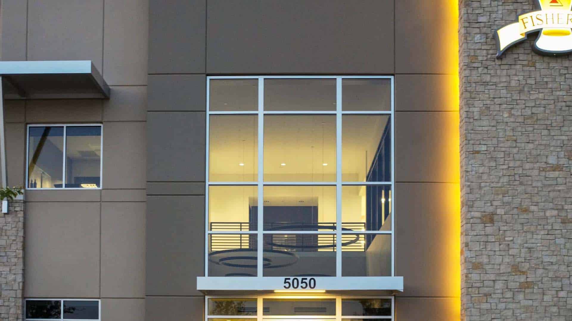 Modern building exterior with large glass windows, illuminated interior lights, and the number 5050 above the entrance. Part of a stone wall and a sign are visible on the right.
