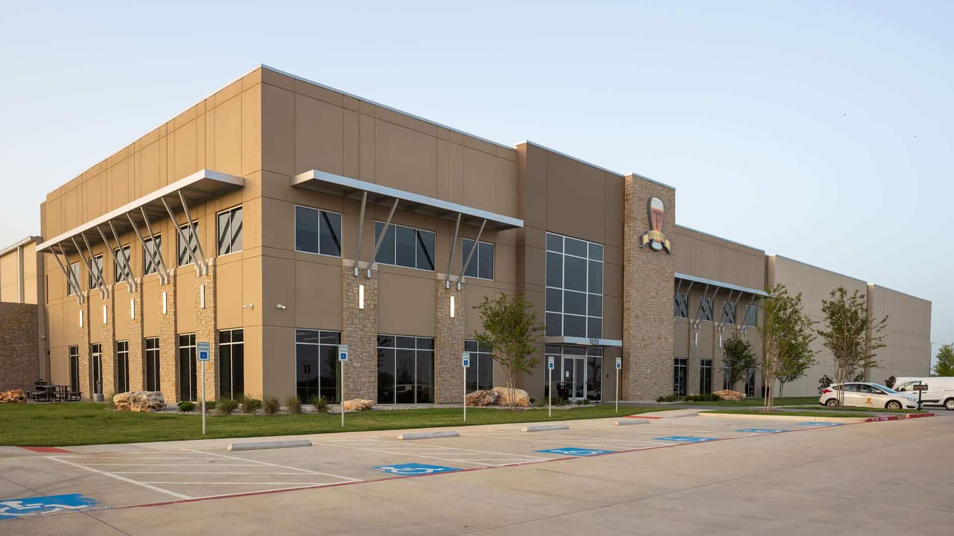 Modern, two-story commercial or industrial building with large windows, a stone facade, accessible parking spaces, and landscaped grounds.