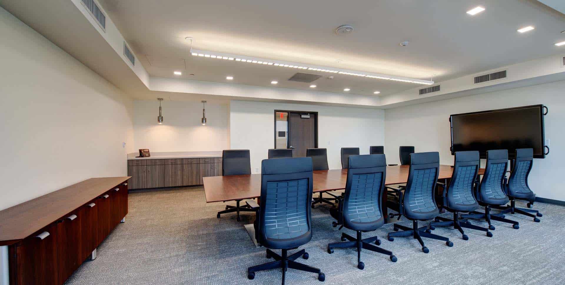 A modern conference room with a long wooden table, ten black chairs, a large screen on the wall, and cabinets along the sides.