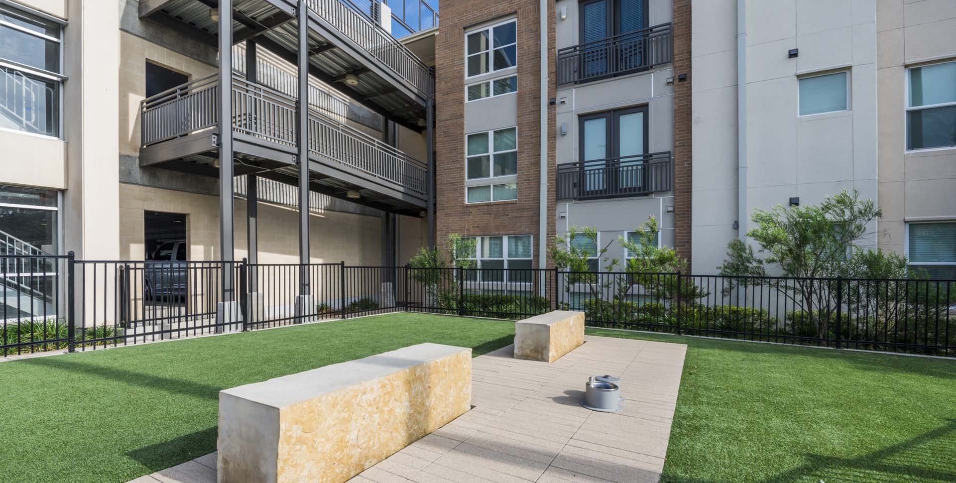 A small outdoor courtyard with artificial grass, two stone benches, a paved walkway, and metal railings, next to a modern apartment building.