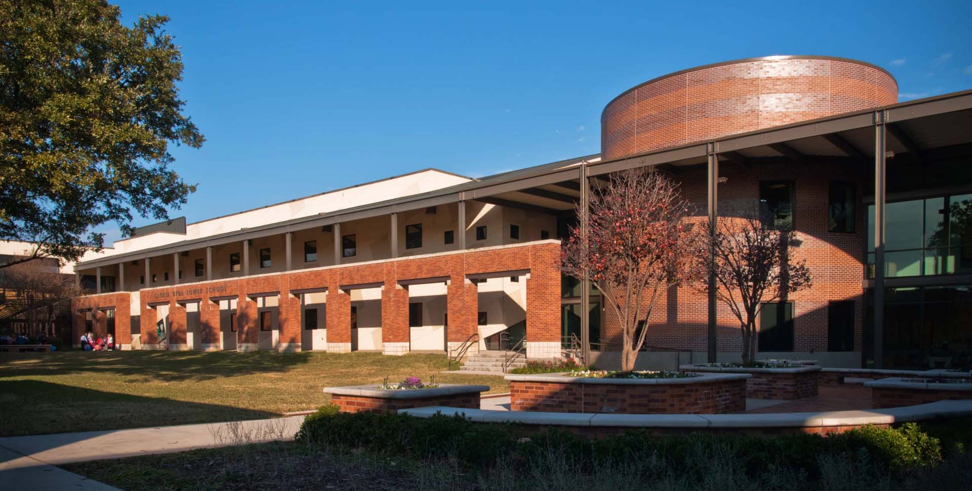 A modern two-story brick building with a circular structure and large windows, surrounded by grass, trees, and a paved walkway under a clear blue sky.