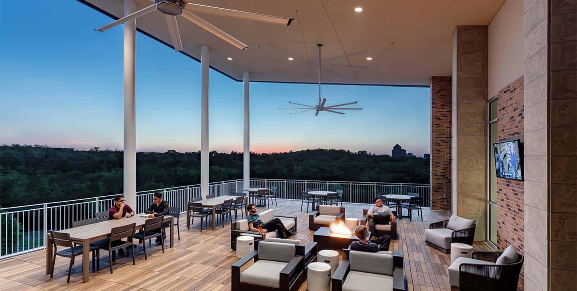 People sit and talk on a spacious outdoor terrace with tables, chairs, and a fire pit at sunset. Ceiling fans and a mounted TV are visible; trees can be seen in the background.