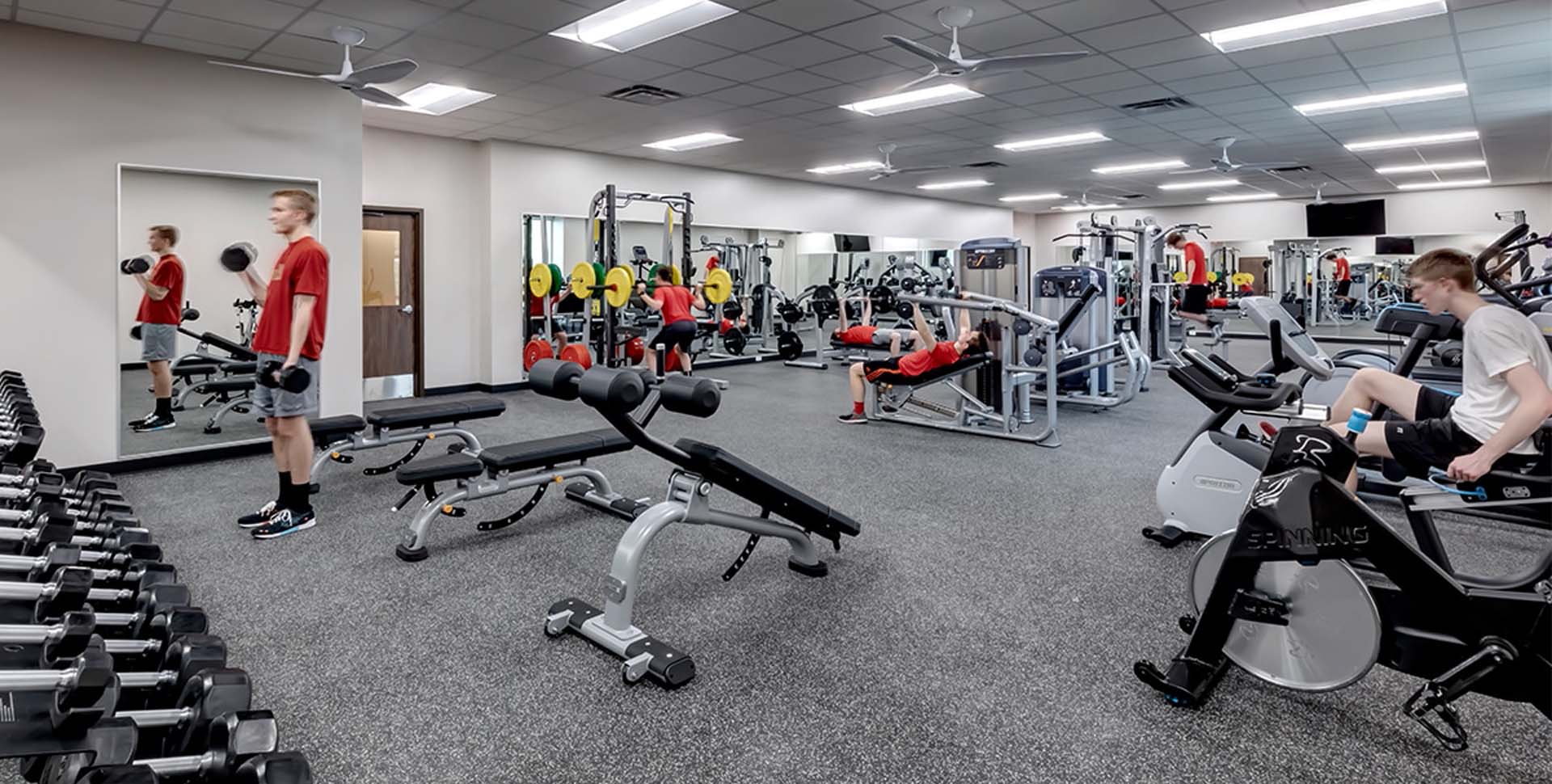 Several people exercise in a spacious gym with various equipment, including weights, benches, cardio machines, and mirrors on the wall.
