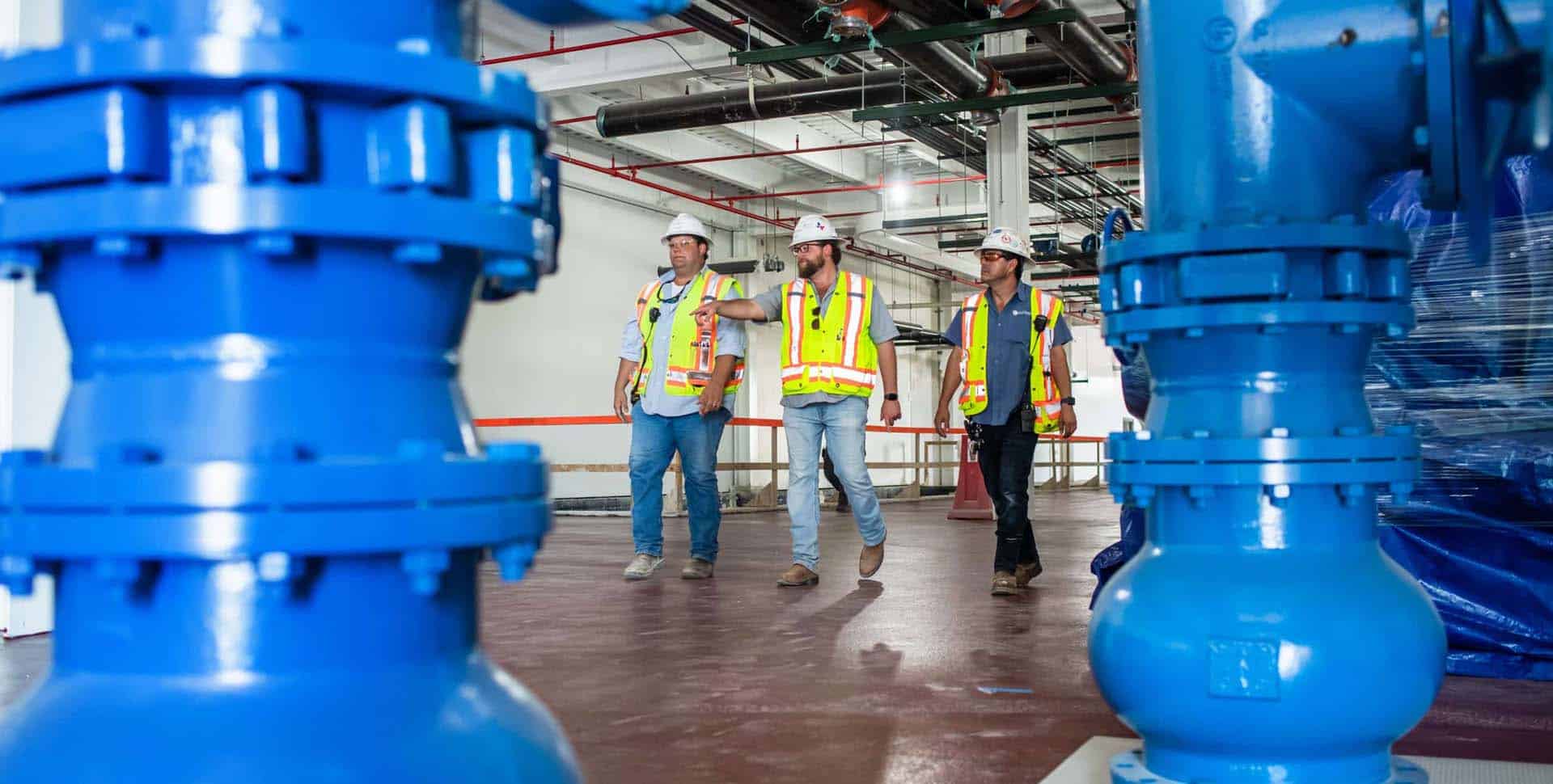Three workers in safety vests and helmets walk through an industrial facility with large blue machinery in the foreground.