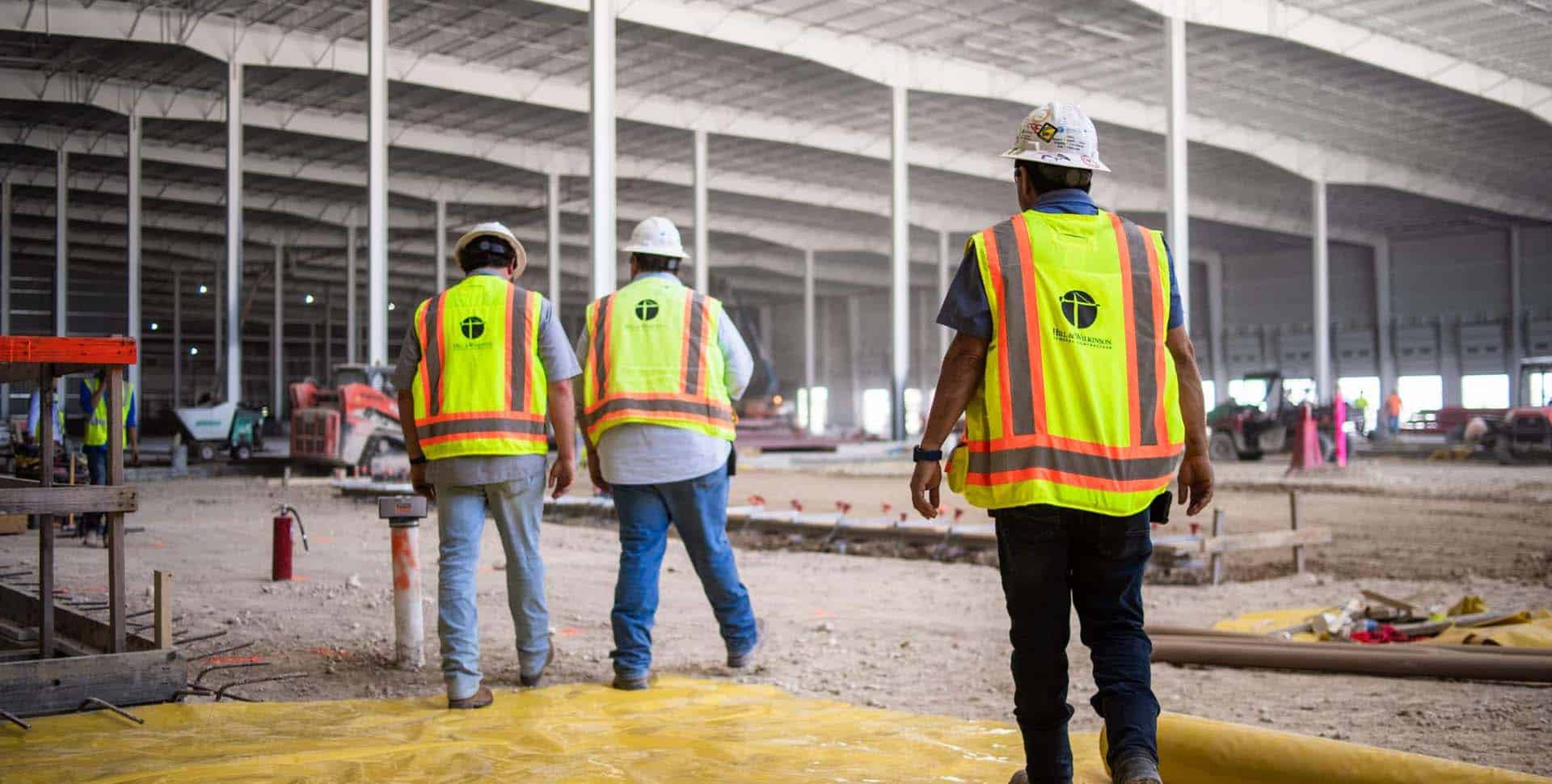 Three construction workers in safety vests and hard hats walk inside a large, unfinished industrial building.
