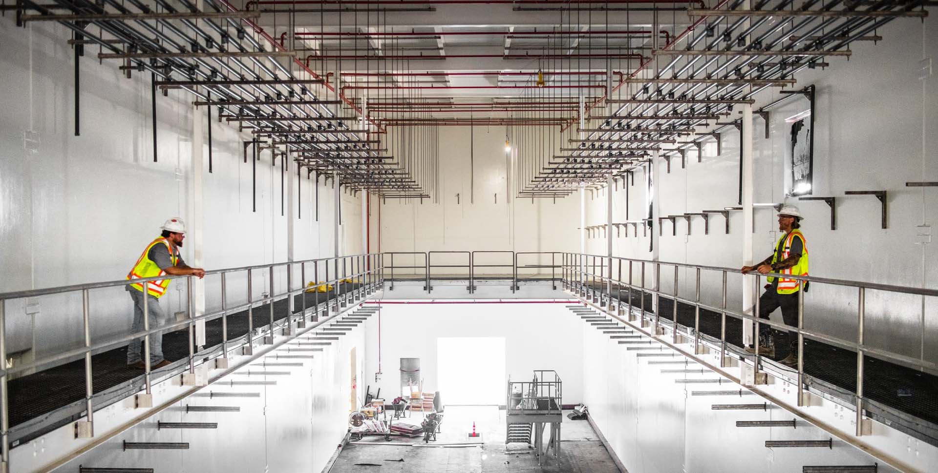 Two construction workers in safety vests and helmets stand on opposite sides of a large, unfinished industrial room with exposed pipes and white walls.