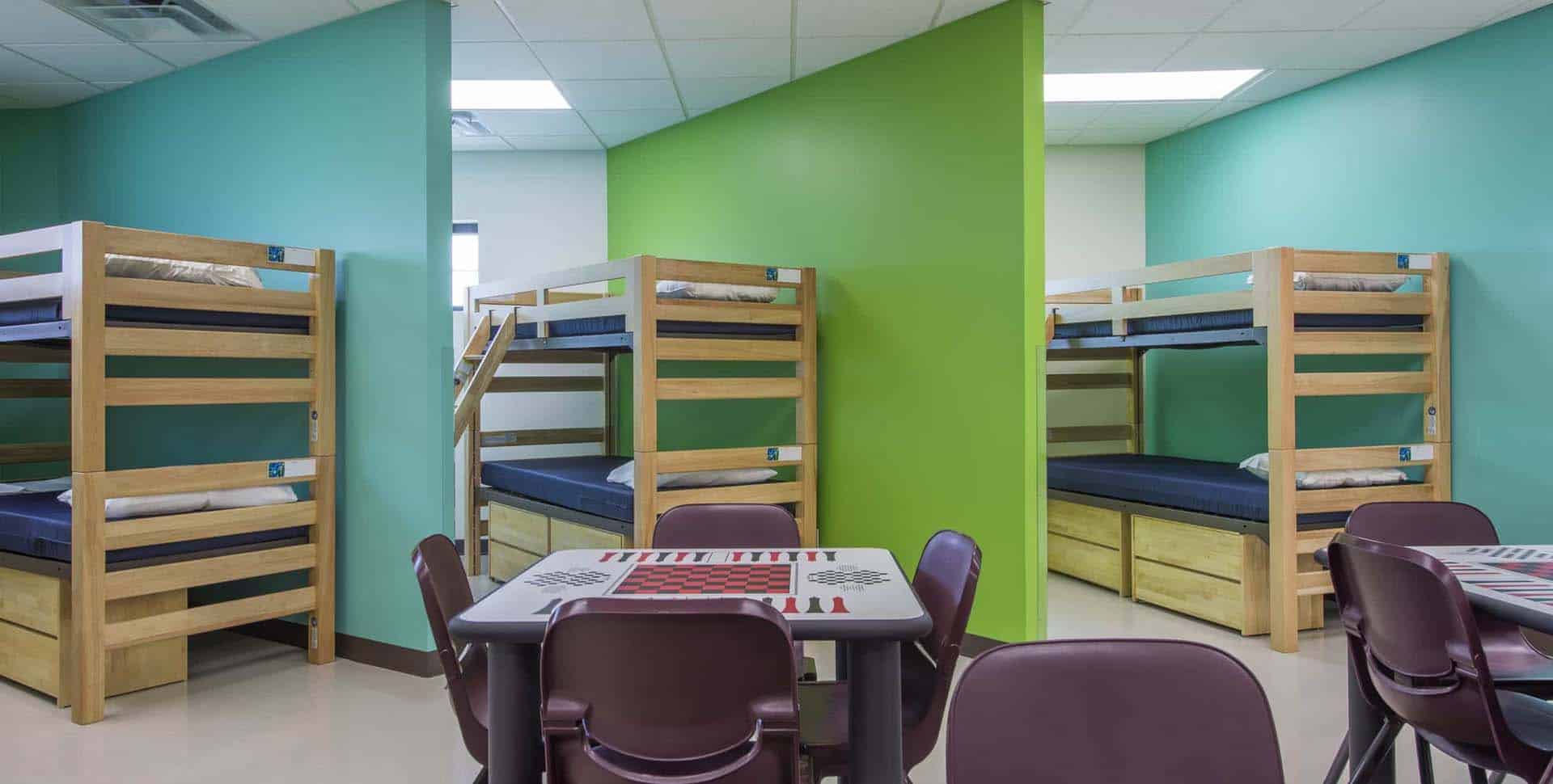 Room with wooden bunk beds against green and teal walls, each bed with bedding. In the foreground, tables and chairs are arranged for seating.