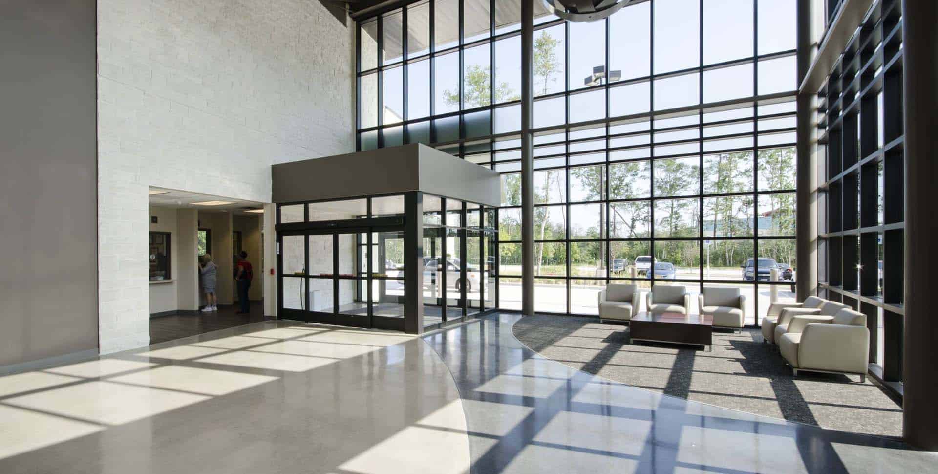 Modern building lobby with large floor-to-ceiling windows, glass entrance doors, seating area with white chairs, and sunlight streaming onto the polished floor.