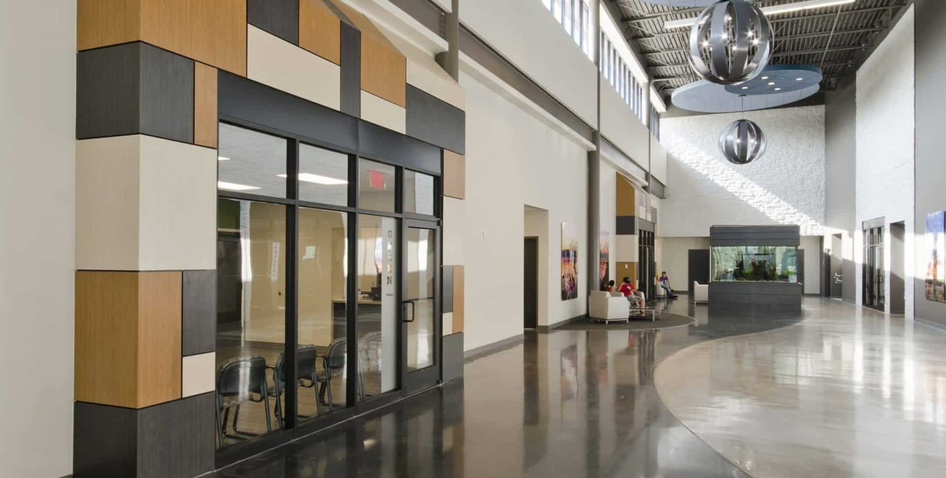 Modern building interior with large windows, geometric wall panels, polished floors, and a waiting area with people sitting near an aquarium.