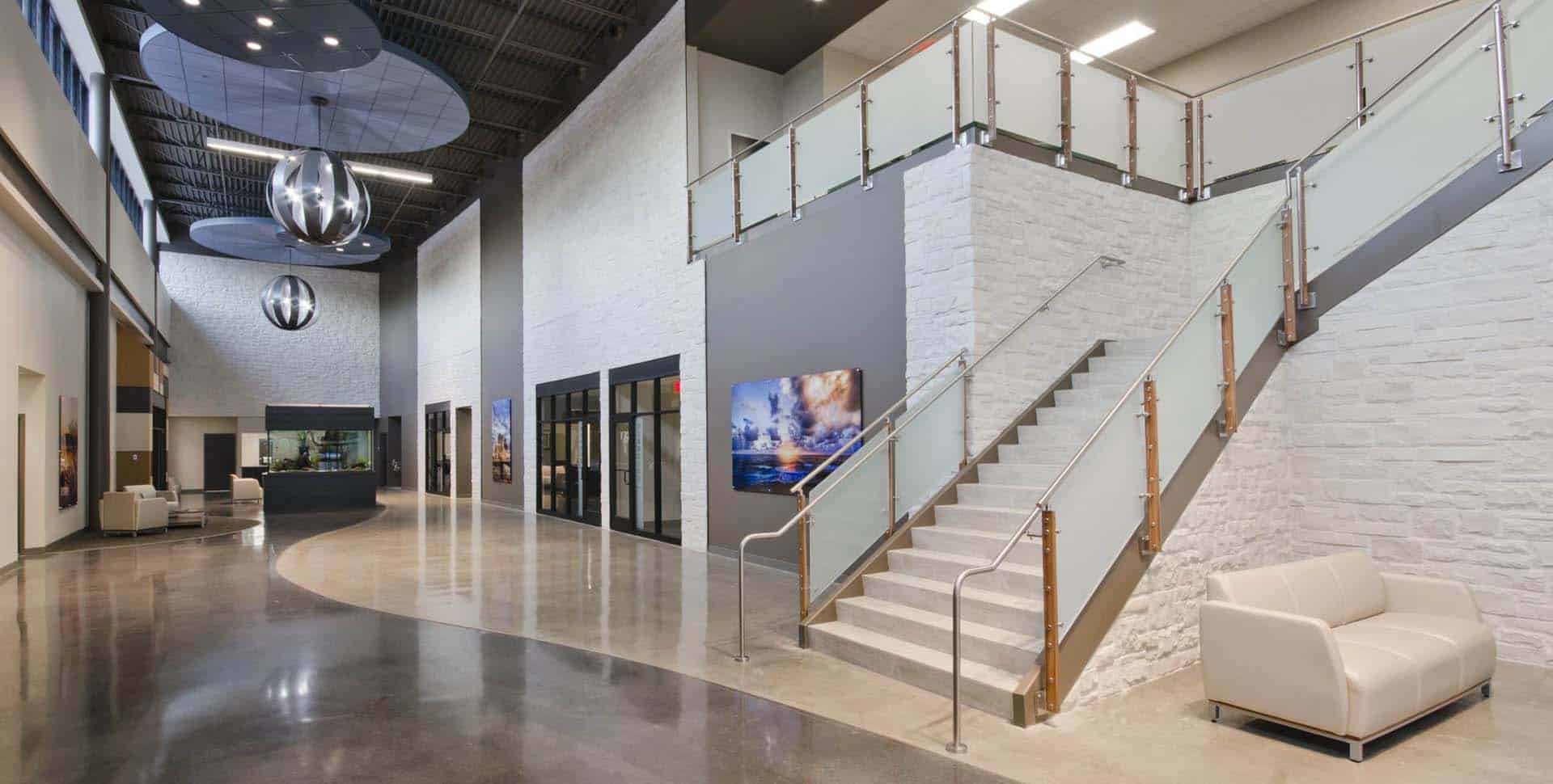 Modern office lobby with white walls, glass railing staircase, polished floors, contemporary lighting fixtures, and seating areas.