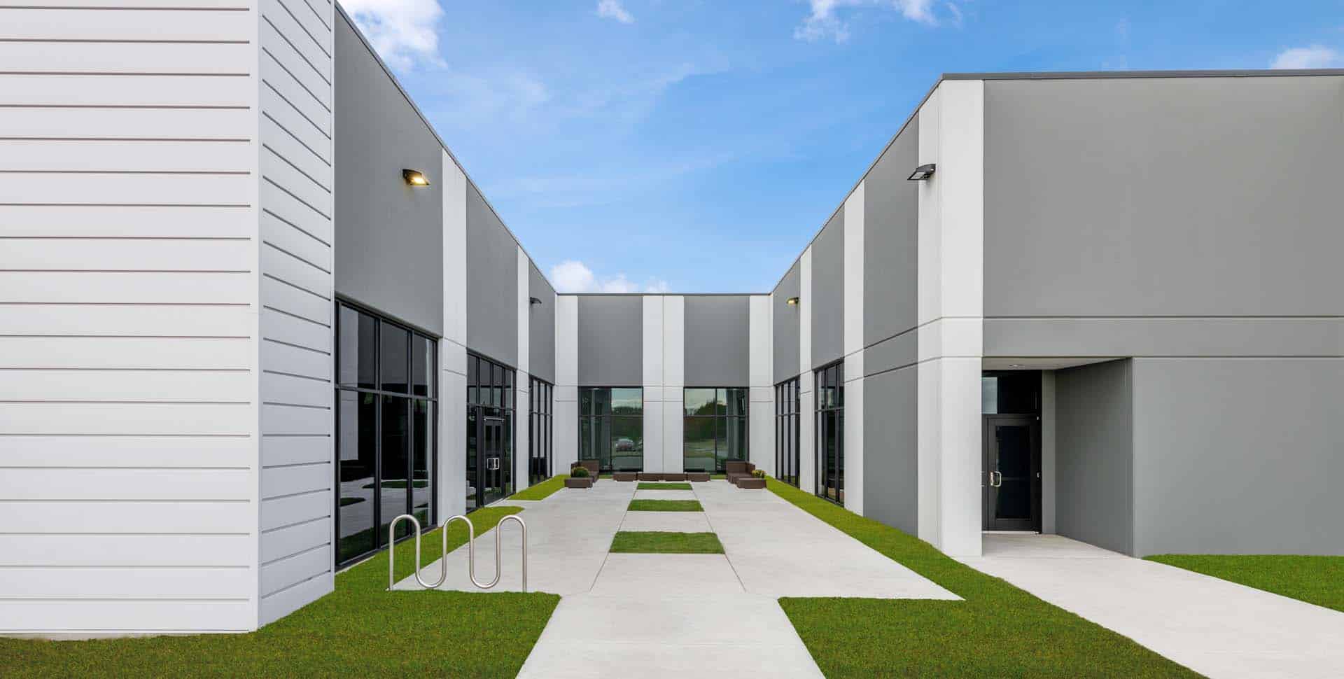 Contemporary commercial building with gray and white exterior, large windows, bike rack, and walkway surrounded by green grass under a blue sky.
