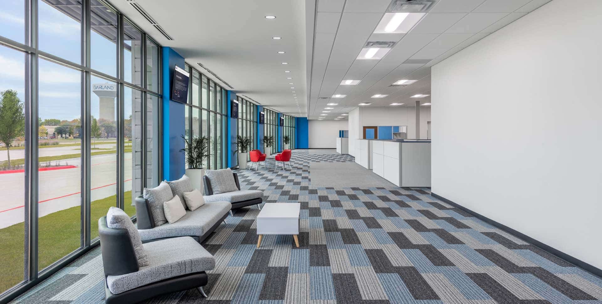Modern office lobby with large windows, patterned carpet, gray seating area with a small table, red chairs in the background, and potted plants along the wall.