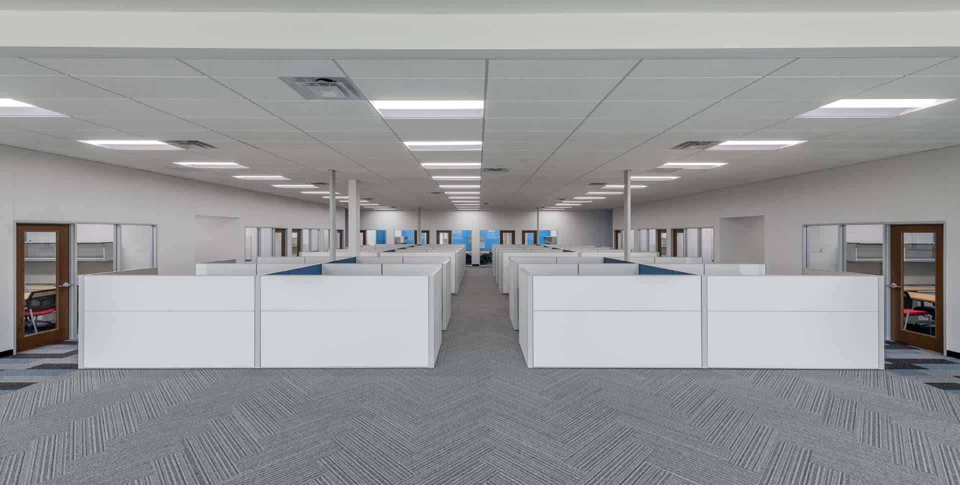 Large open-plan office with rows of white cubicles, gray carpeted floor, and fluorescent ceiling lights; empty chairs visible within the cubicles.