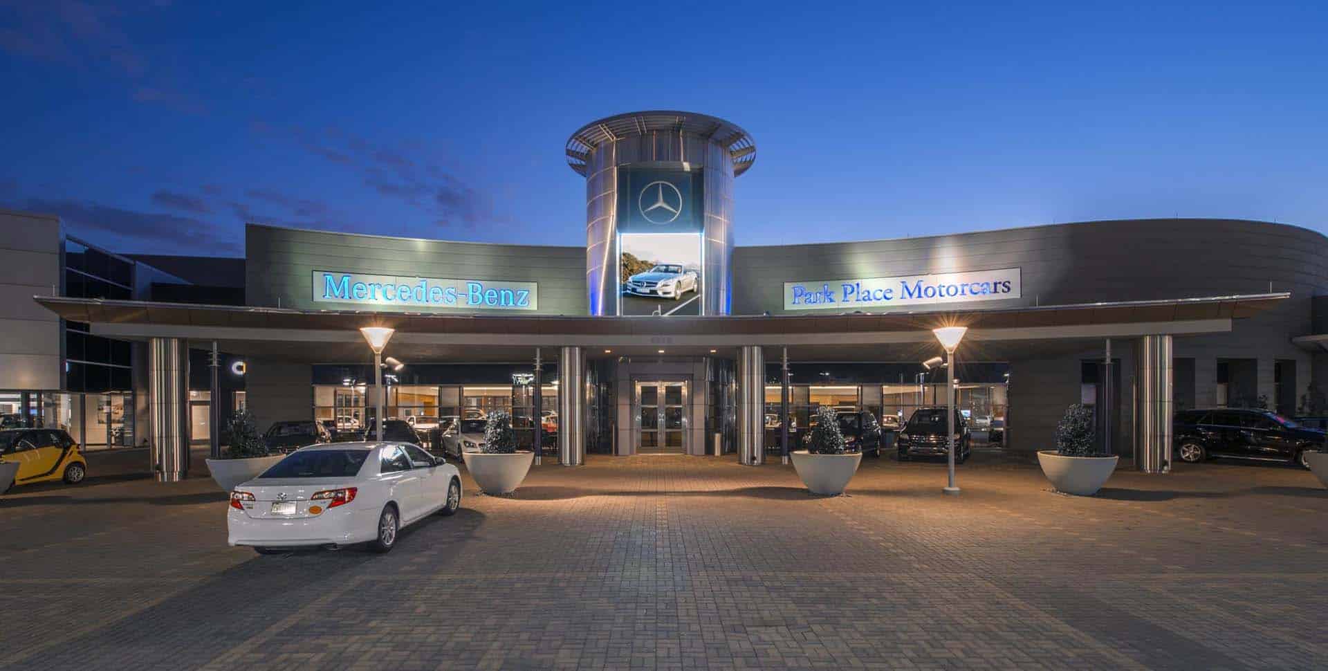 Front entrance of a Mercedes-Benz Park Place Motorcars dealership at dusk, with cars parked outside and illuminated signage above the doors.