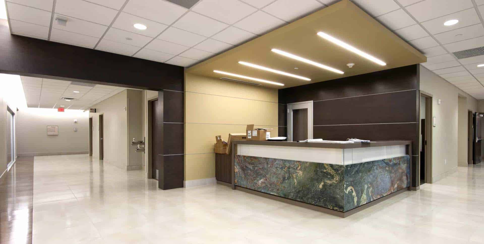 A modern, empty hospital reception area with a marble-front desk, beige floor tiles, and overhead lighting, adjacent to a clean hallway.