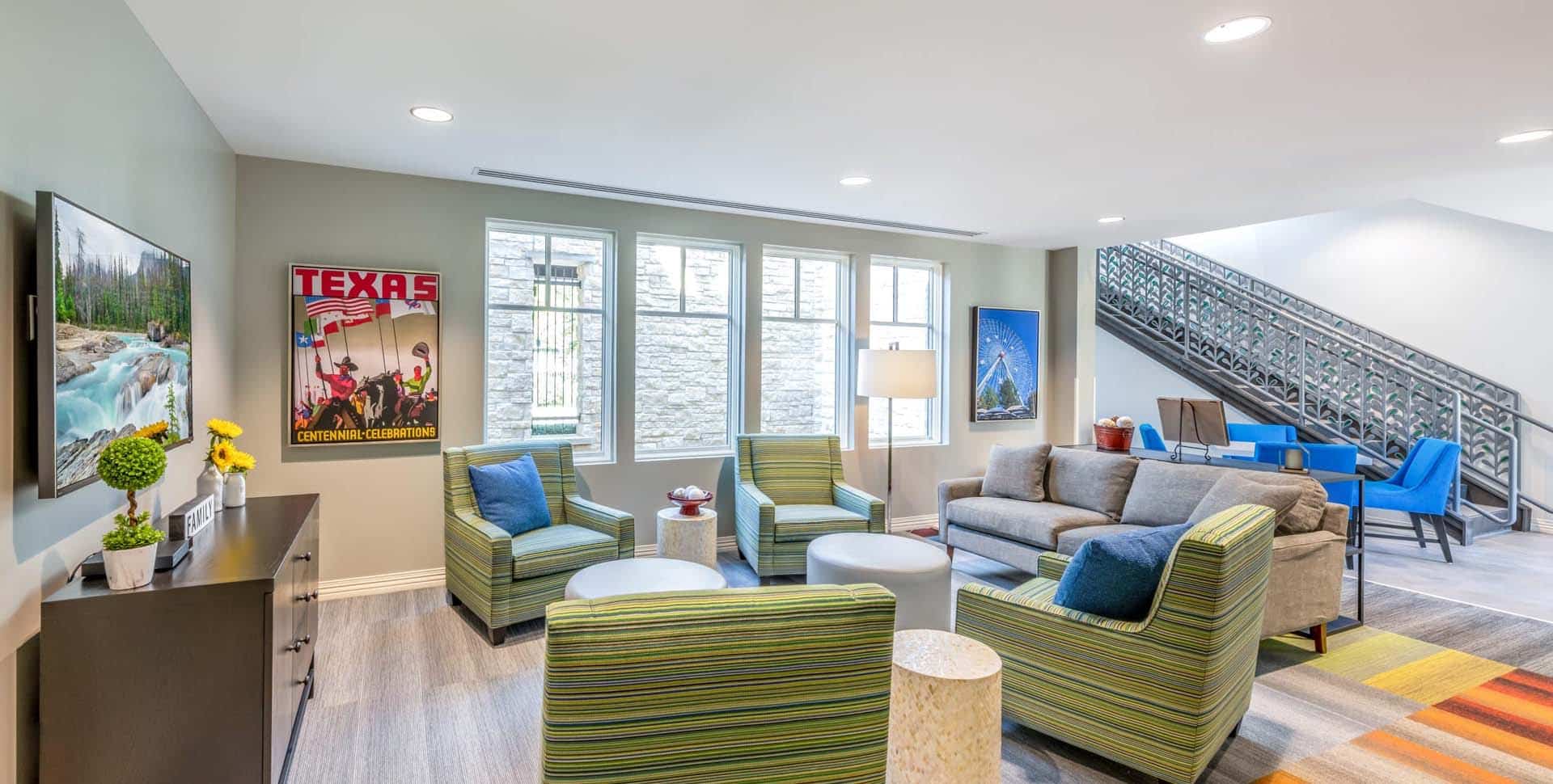 Modern lounge area with green-striped chairs, a gray sofa, colorful rug, wall art, and a staircase leading to an upper level. Large windows let in natural light.