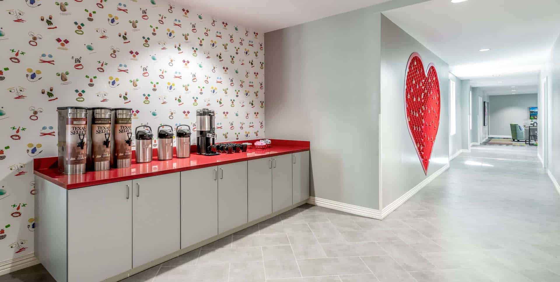 A coffee station with drink dispensers and cups on a red countertop next to a hallway with a large red heart artwork on the wall.