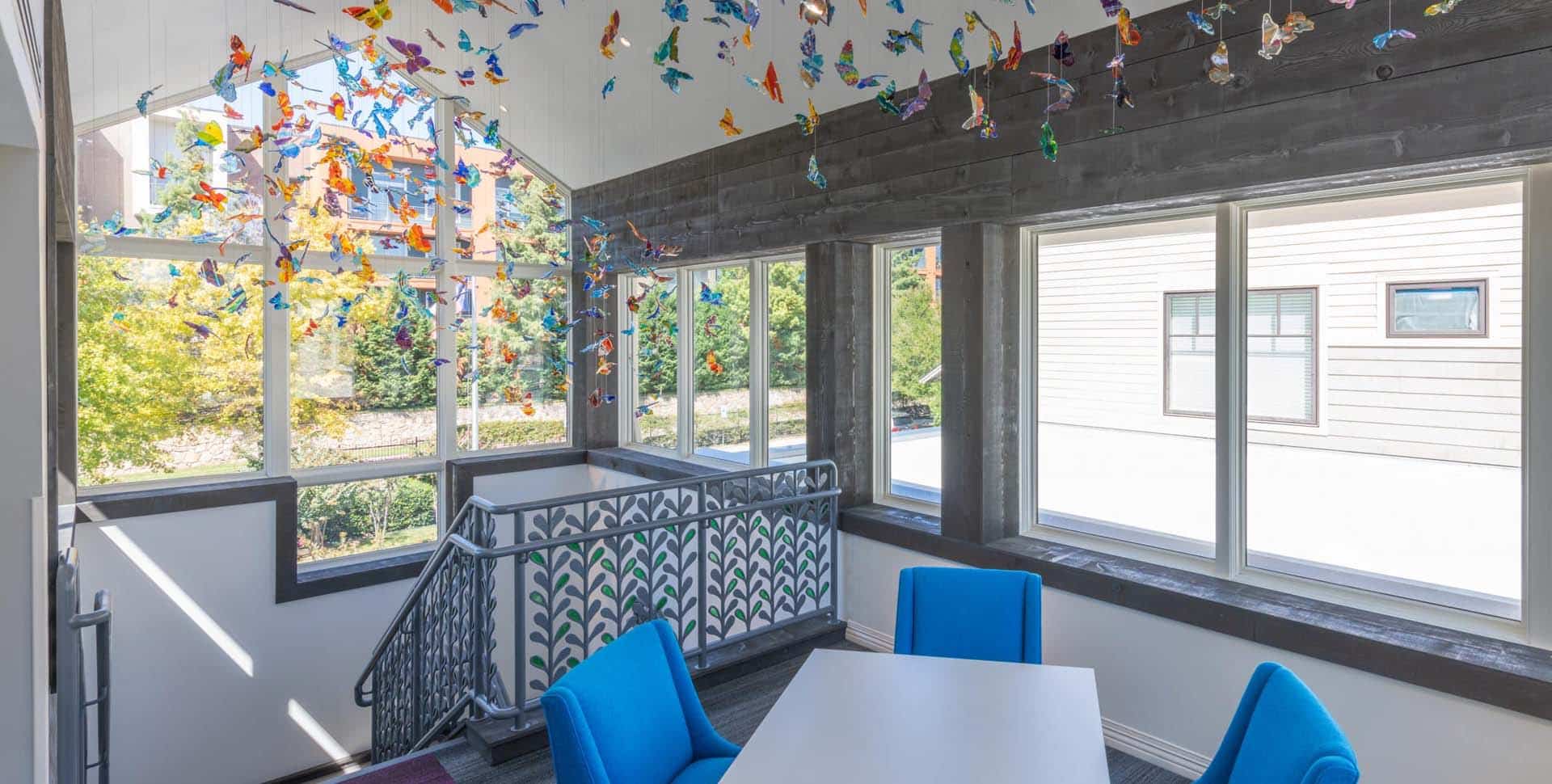 A small meeting area with blue chairs and a white table near windows, featuring colorful hanging butterfly decorations from the ceiling.