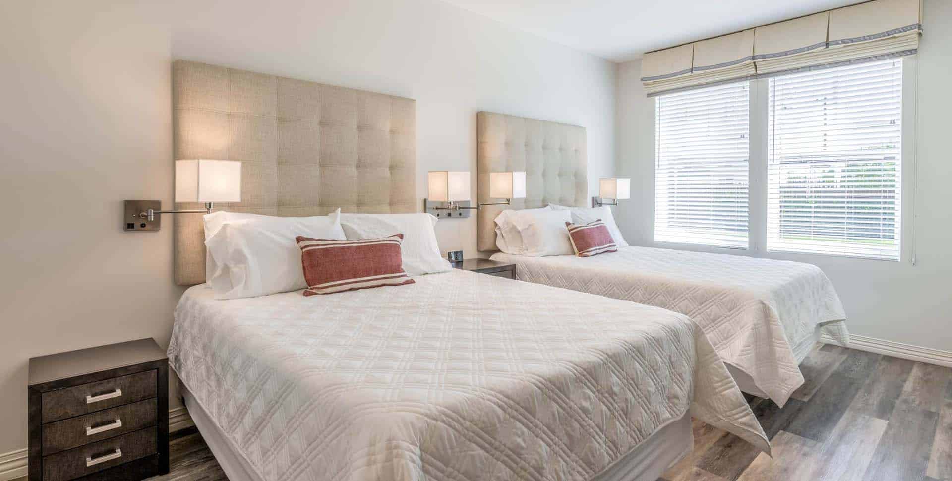 A bright bedroom with two double beds, each with a tufted headboard, white bedding, and red accent pillows, next to a window with blinds and two bedside tables.