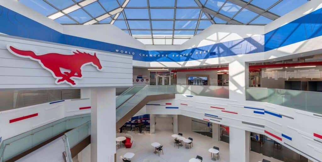 Modern interior of a building with a glass ceiling, red mustang logo, "World Changers Shaped Here" sign, tables, and blue and red accents on walls and railings.