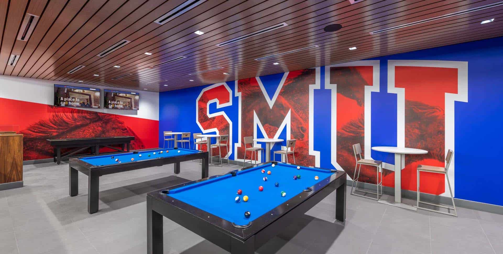 A modern game room with two pool tables, high tables with chairs, TVs, and a bold mural featuring large SMU letters on the wall.
