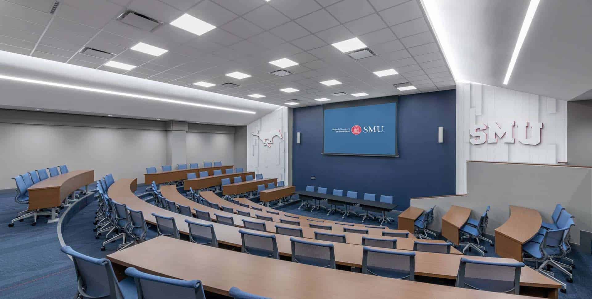 A modern lecture hall with curved rows of wooden desks and blue chairs facing a large screen displaying the SMU logo and branding.