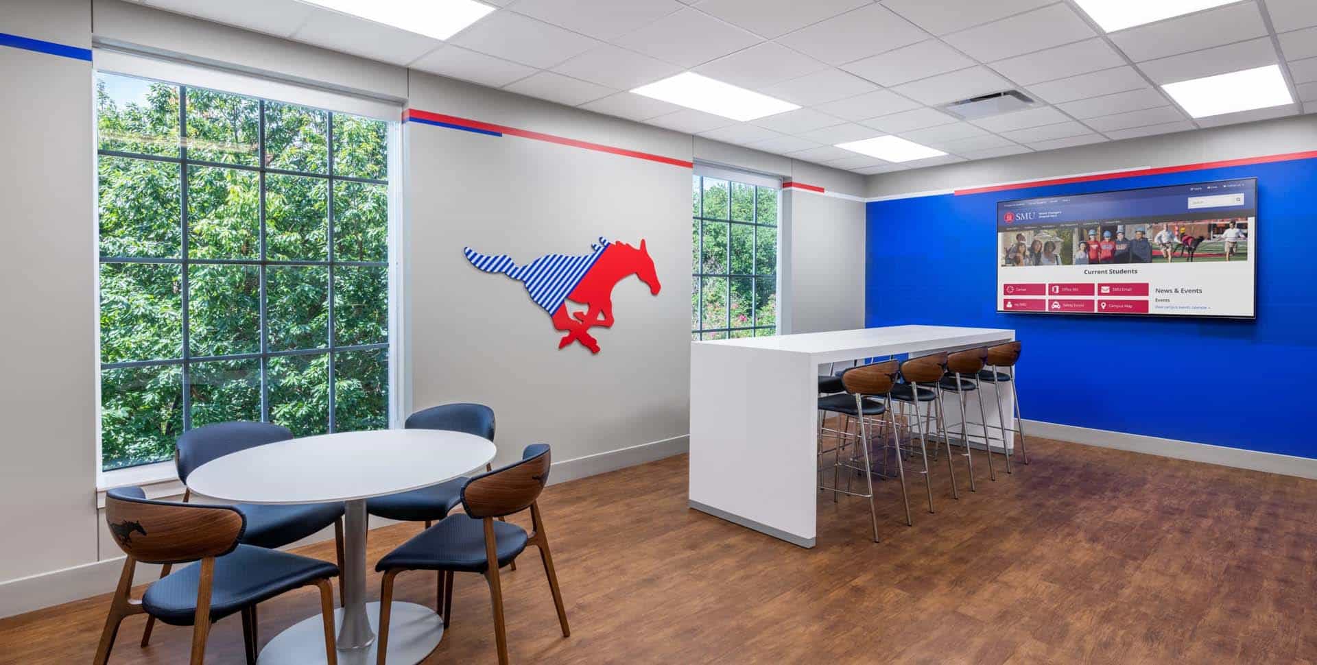 A modern lounge area with round tables, chairs, a high counter, wall art of a horse, large windows, and a wall-mounted screen displaying a website.