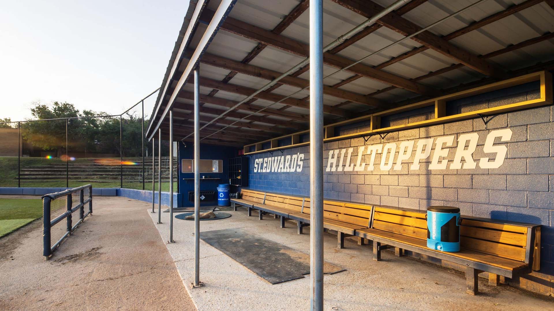 Empty baseball dugout with wooden benches, a blue trash can, and "St. Edward's Hilltoppers" written on a blue cinder block wall.