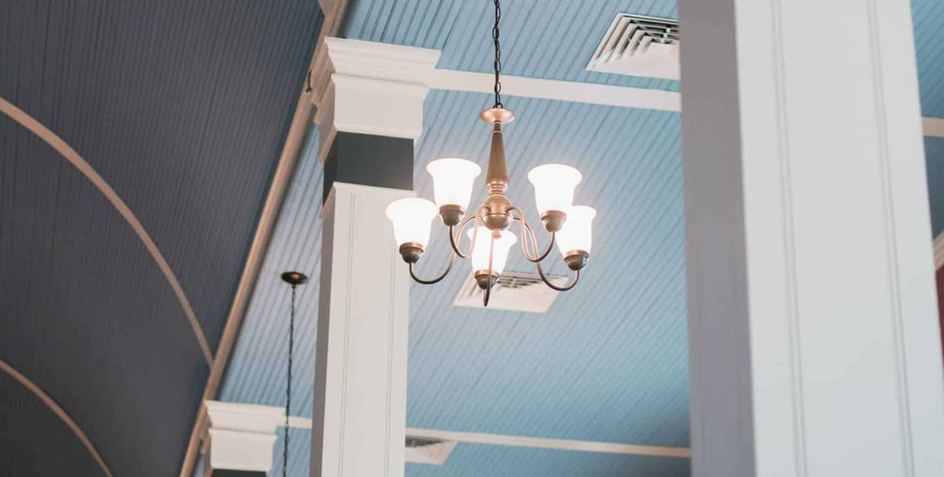 A vintage brass chandelier with five lit bulbs hangs from a high ceiling with blue paneling and white trim.