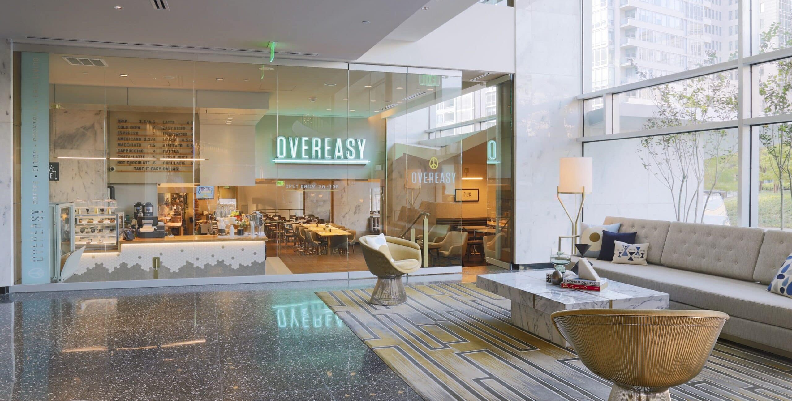 A modern café with large glass windows and a neon “Overeasy” sign above the entrance. Inside are wooden tables, chairs, and a counter with coffee machines and pastries. In front of the café, a lounge area features a beige sofa with patterned pillows, a marble coffee table, a gold wire‑frame chair, and a geometric rug. Large windows fill the space with natural light.
